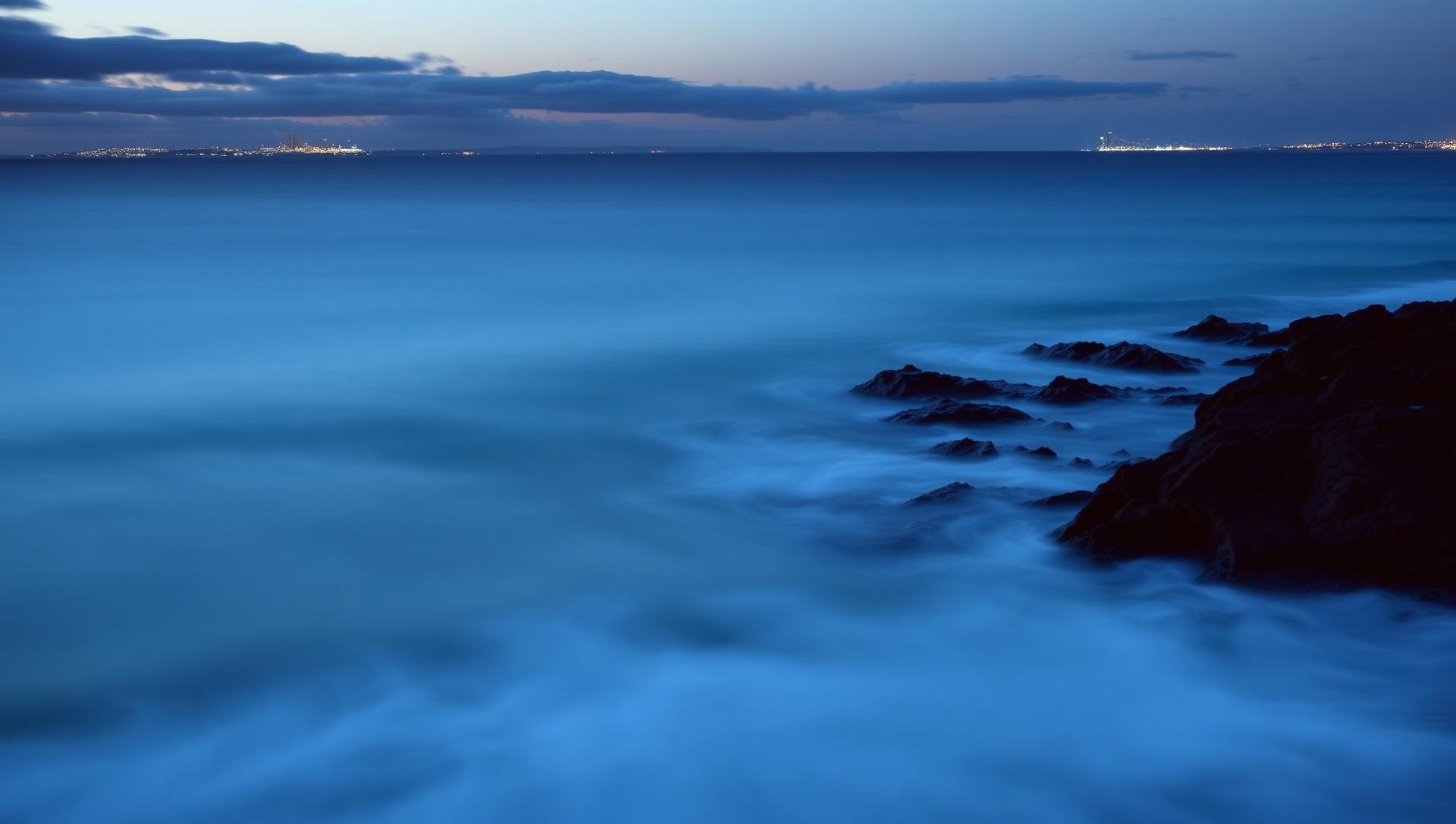 Gold Coast coastline at dusk, deep blue Pacific meeting basalt headland rocks with distant city lights on the horizon